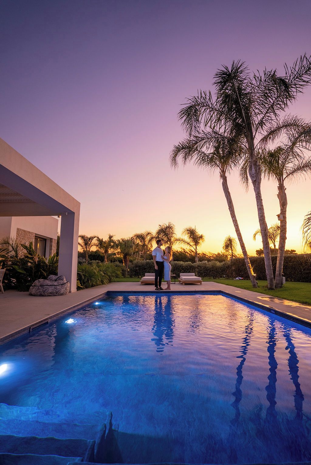 A swimming pool with a sunset in the background, surrounded by palm trees and a modern building.