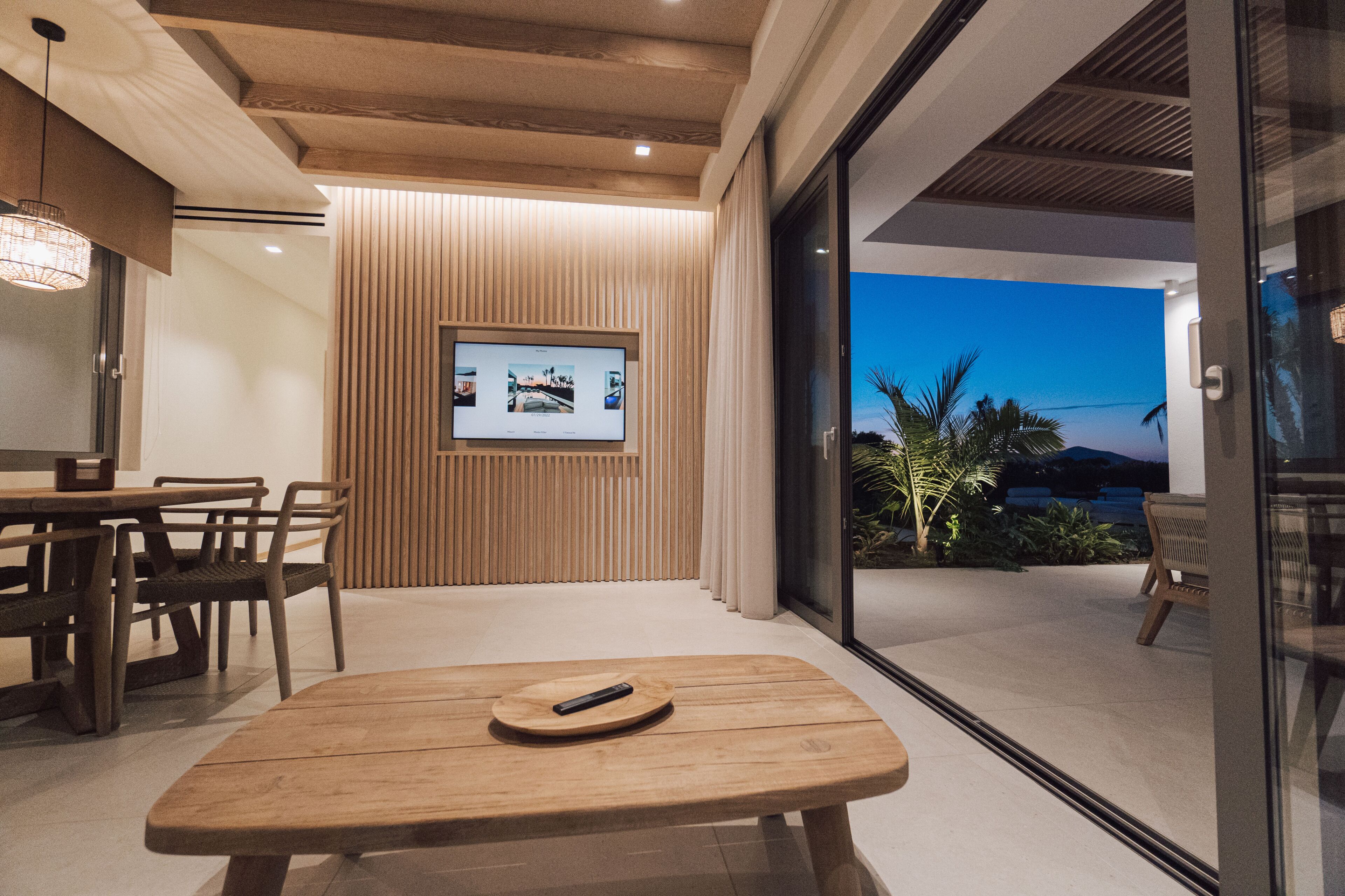 A modern interior with a wooden table and chairs, a television mounted on a wall, and a view of the outdoors through a glass door.
