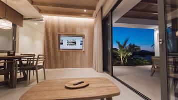 A modern interior with a wooden table and chairs, a television mounted on a wall, and a view of the outdoors through a glass door.