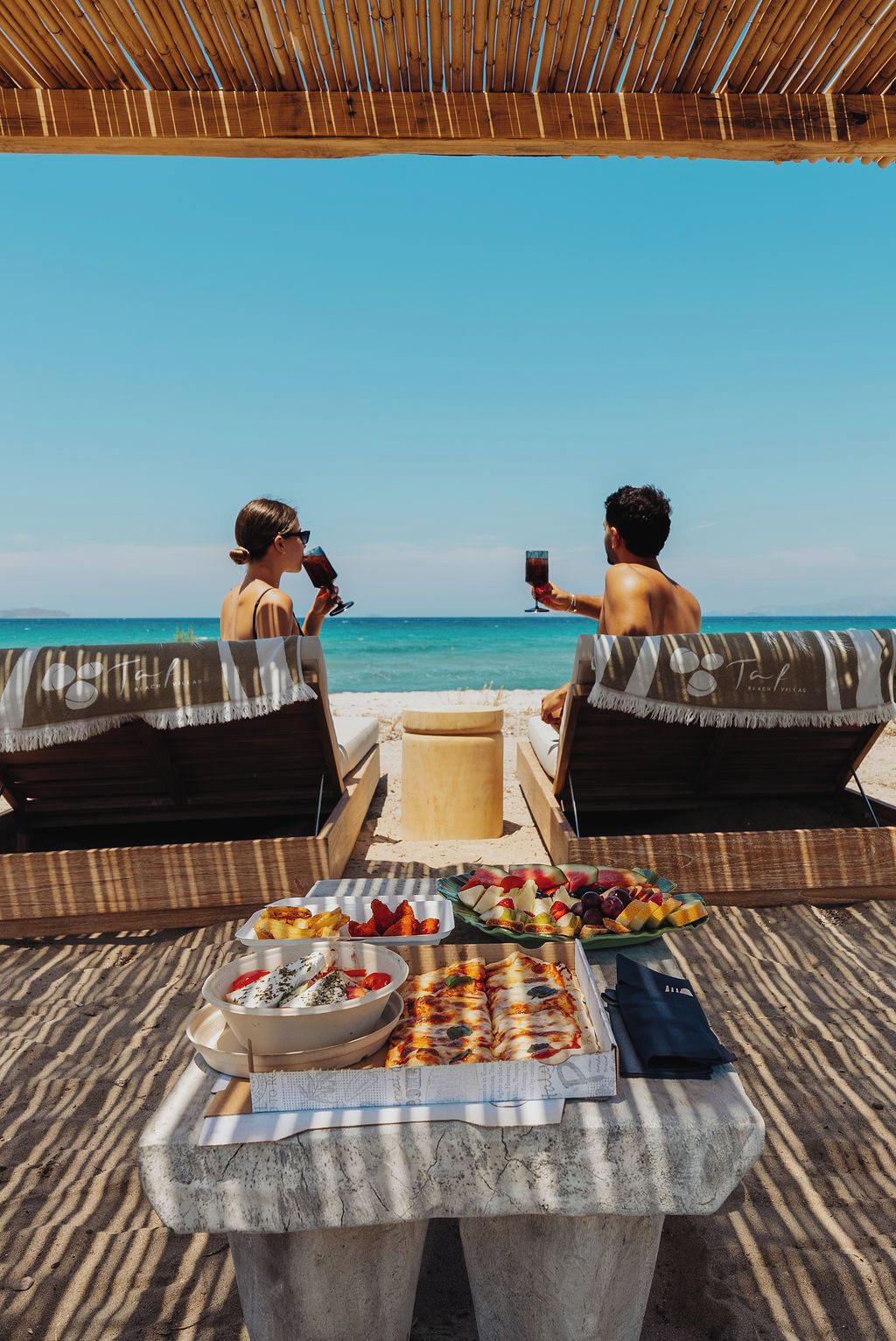 Escena frente al mar con dos sillas de playa mirando al océano, una mesa con comida y bebidas, y una vista despejada del mar.