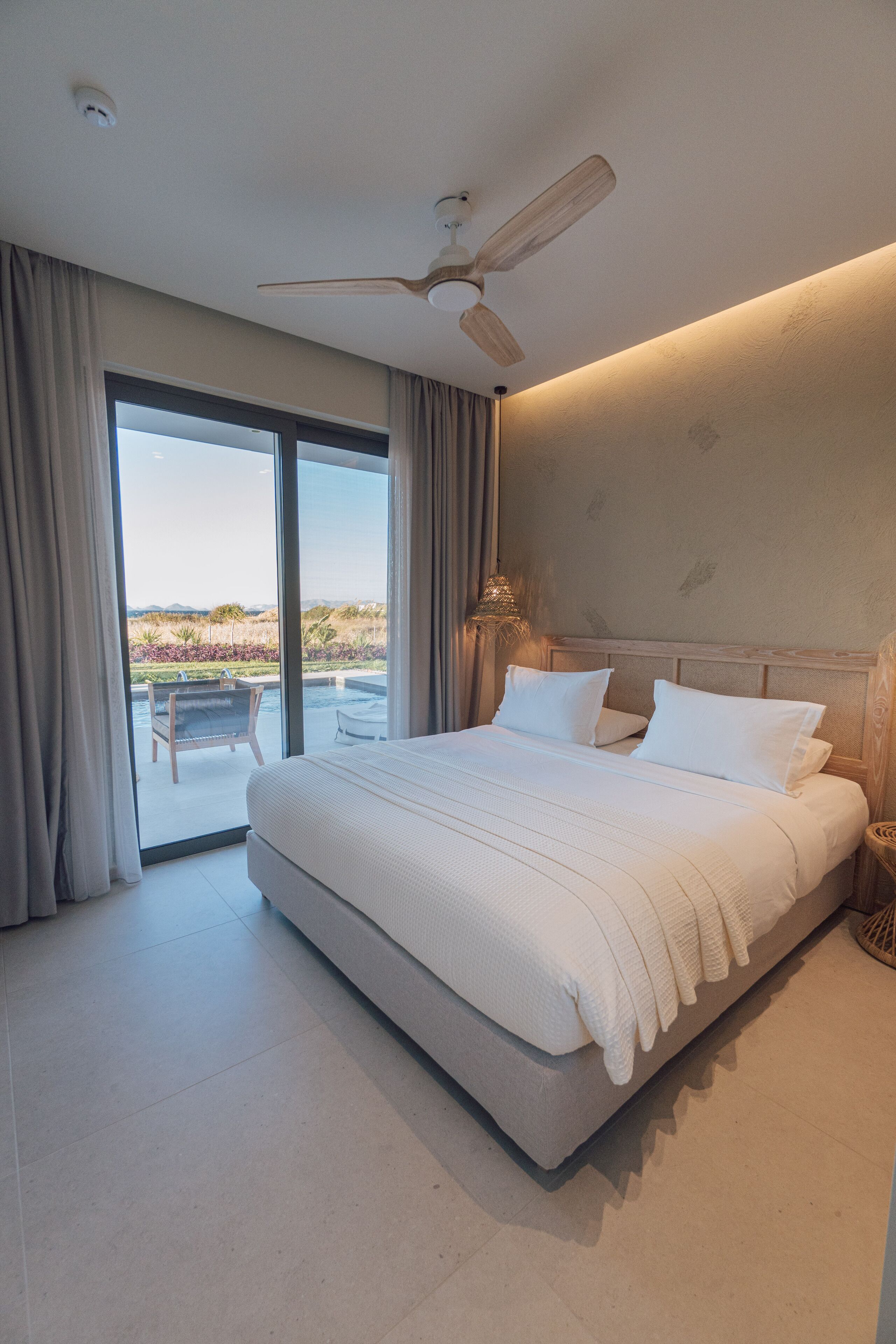 A modern bedroom with a large bed, a ceiling fan, and a view of a landscaped area through a sliding glass door.
