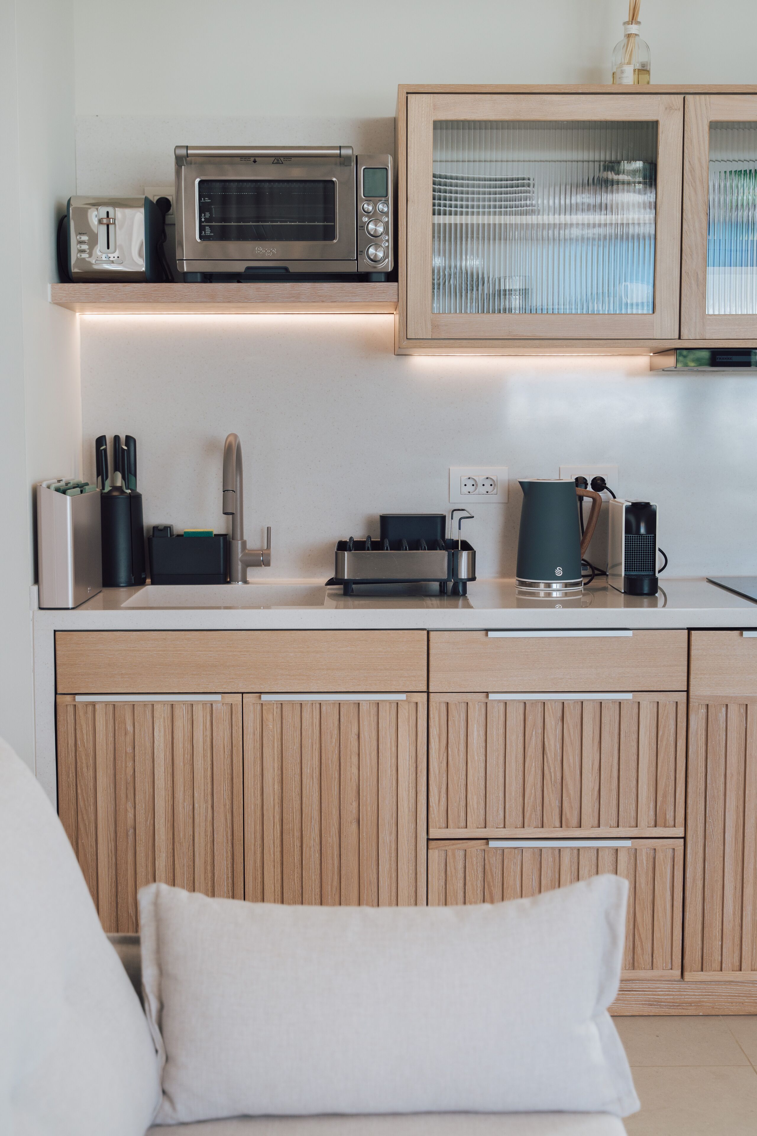 A modern kitchen with a built-in microwave, toaster, and coffee maker. There is a wooden cabinet with a glass door above the counter.