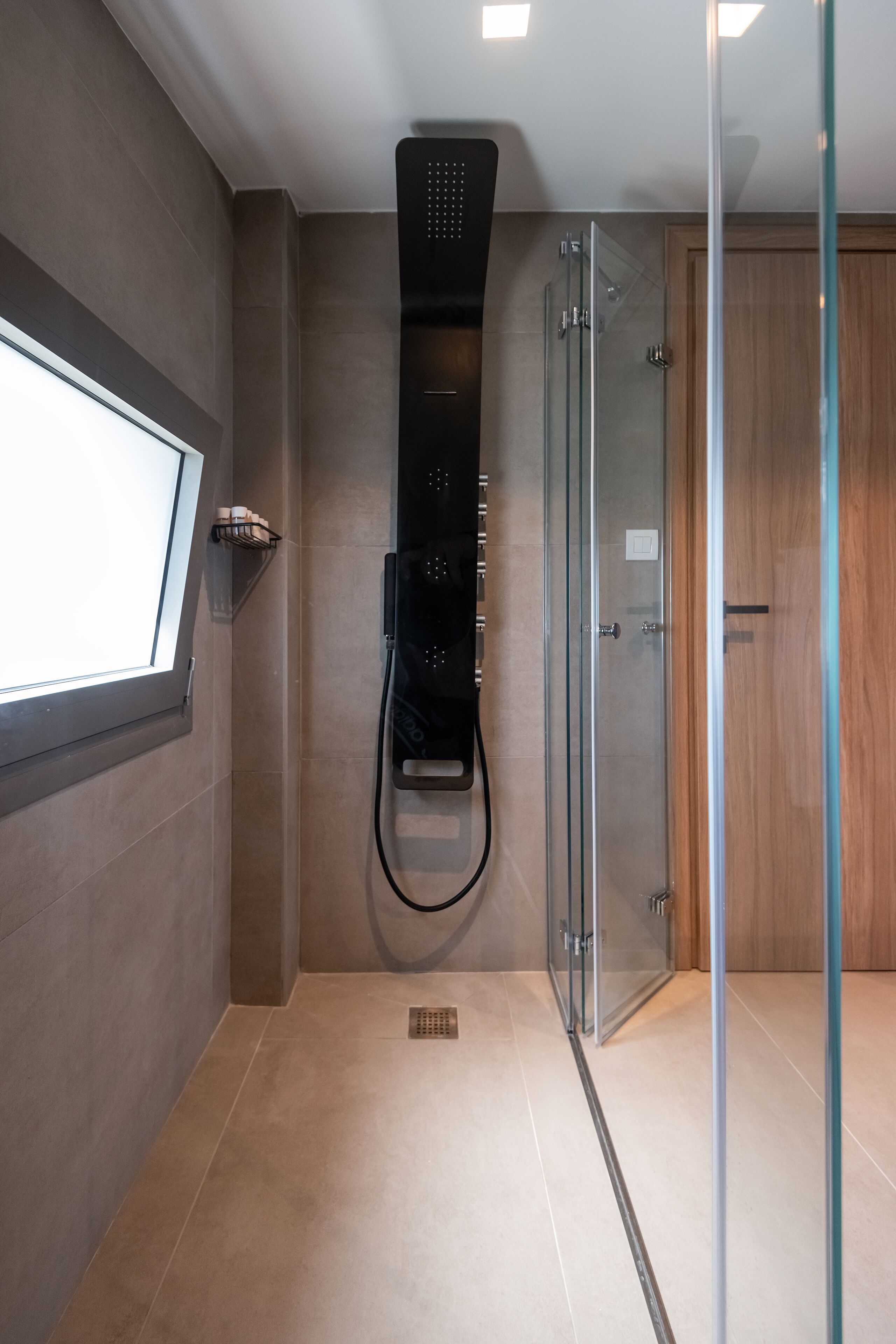 A modern bathroom with a glass shower enclosure, a built-in towel rack, and a wooden door.