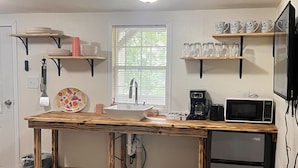 Una cocina con una mesa de madera, un fregadero, un microondas y un refrigerador. Hay estantes sobre el fregadero y una ventana con persianas.