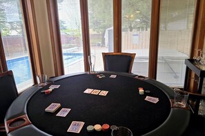 Mesa redonda com cartas de baralho e fichas de poker, rodeada por cadeiras, com grandes janelas ao fundo.