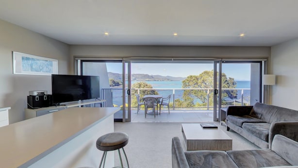 Un salon moderne comprenant un canapé, une table basse et un téléviseur. Il dispose d’un balcon offrant une vue sur la mer et les montagnes.