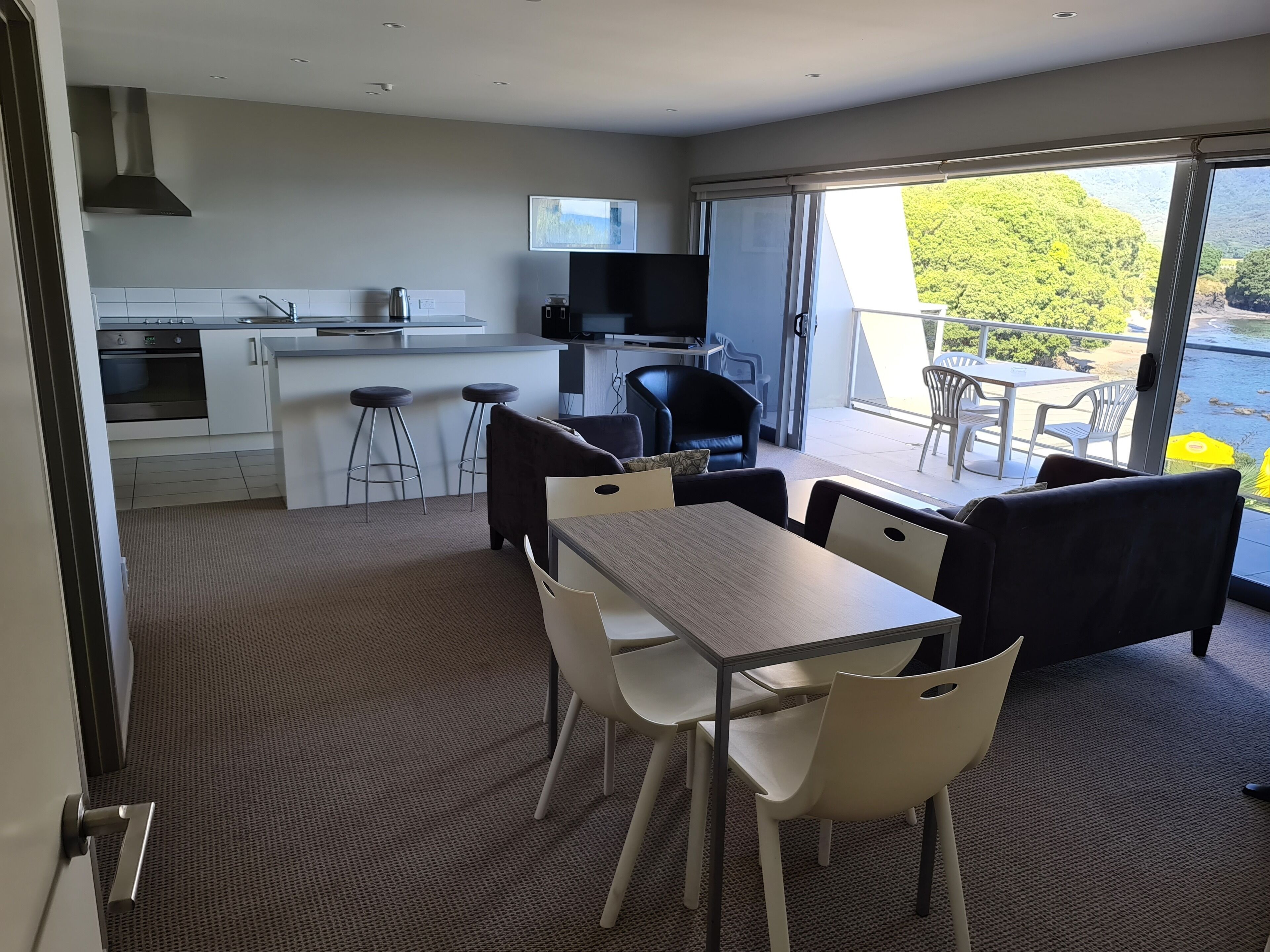A modern kitchen with white cabinets, a stainless steel range hood, and a built-in oven. There is a dining area with a wooden table and white chairs, and a balcony with a view of greenery.