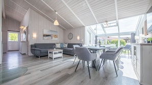 A spacious living area with a dining table, grey chairs, a sofa, and a kitchen area with cabinets and a window. - Baltic View - Komfortables, Sehr Strandnahes Ferienhaus mit Whirlpool und Sauna (Brodersby)
