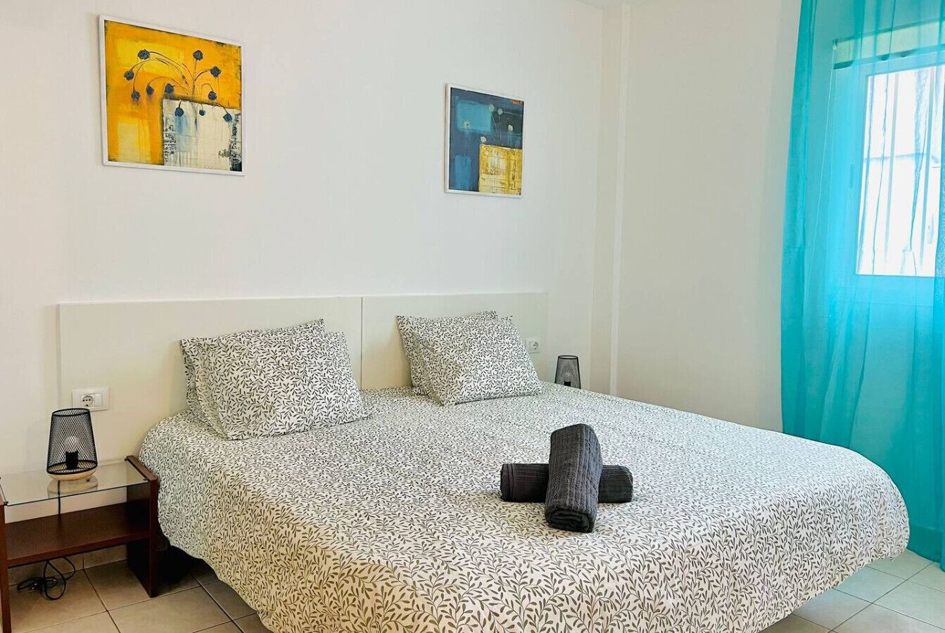 A bedroom with a patterned bedspread, two pillows, a bedside table with a lamp, and artwork on the walls.