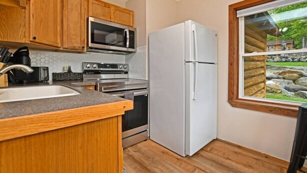 A kitchen with wooden cabinets, a stainless steel microwave, a white refrigerator, and a window with a view of a stone wall and greenery. - Walk to Beach - Close to Town - Pet Friendly Cabin (Lake George)