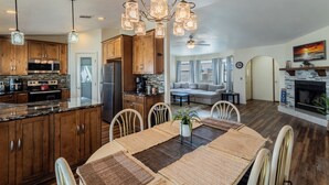 Een ruime keuken met houten kastjes, een granieten aanrechtblad en een centraal eiland. Er is een zithoek met een grote tafel en stoelen, en een woonkamer met een bank en open haard.