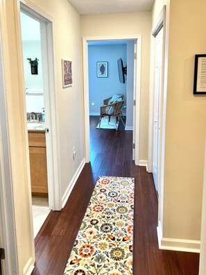 Um corredor com tapete estampado com flores, piso de madeira e uma porta que dá para um quarto com parede azul e uma cadeira.