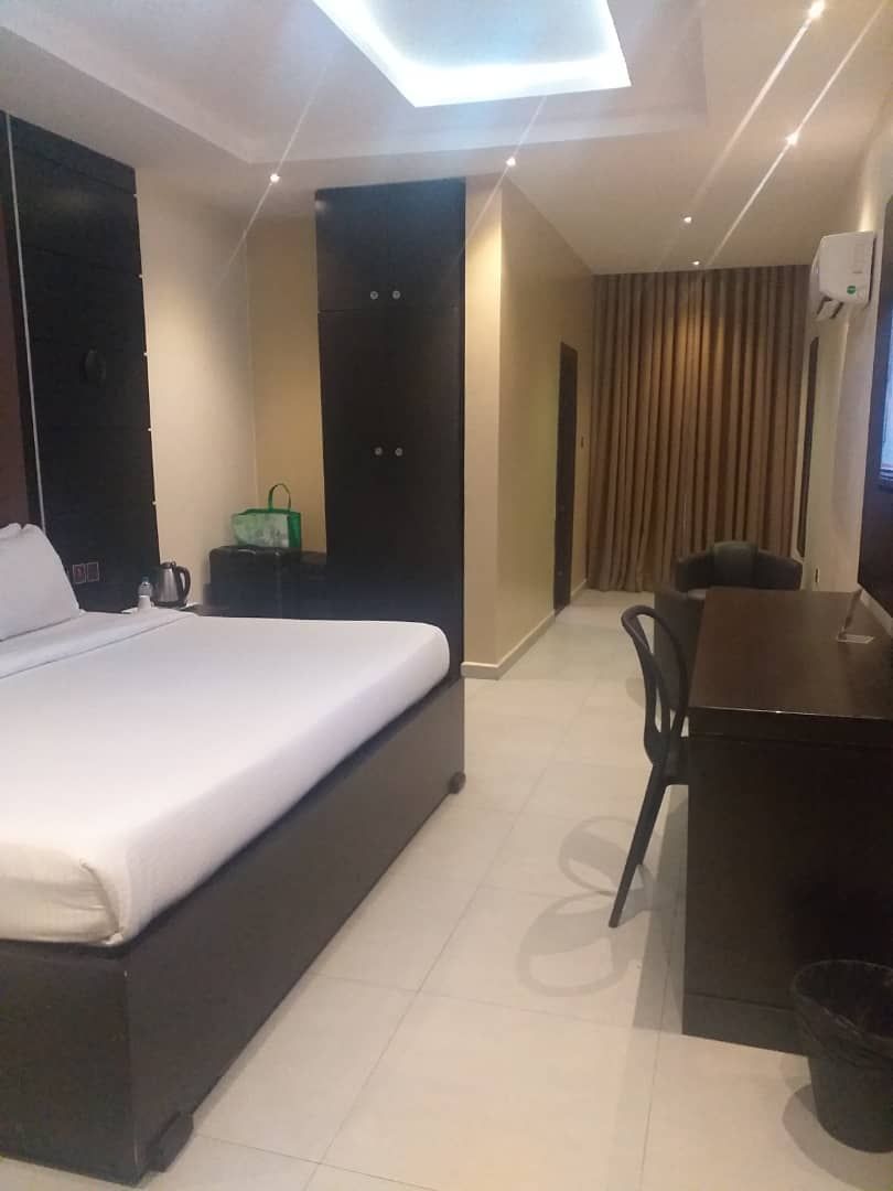 Quarto de hotel com cama, escrivaninha e cadeira. Há uma janela com cortinas, uma luminária no teto e um ar-condicionado fixado na parede.