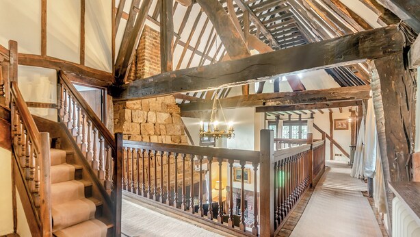 A rustic interior with exposed wooden beams, a stone wall, a wooden staircase, and a hanging chandelier. - 6 bedroom accommodation in East Grinstead (East Grinstead)