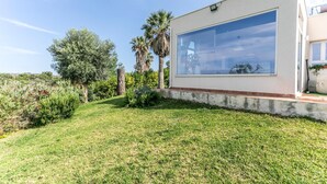 Une maison moderne dotée d’une grande baie vitrée, entourée d’un jardin bien entretenu où poussent différentes espèces de plantes et où s’étend une pelouse.