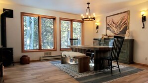 A dining area with a wooden table, chairs, a bench, and a chandelier. There are large windows with a view of trees.