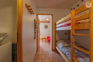 Ein schmaler Raum mit einem Holzbett, einem kleinen Tisch und einer Tür zu einem anderen Raum.