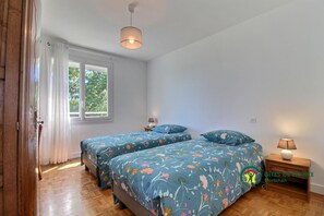 A bedroom with two beds, a window, a wooden door, and a nightstand with a lamp. - Chez Rosalie - Ploemeur (PLOEMEUR)