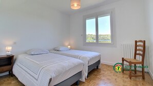 A bedroom with two beds, a wooden chair, a window with a view, and a hanging light fixture.