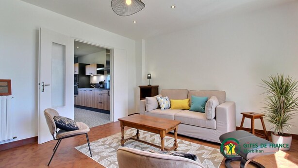 A modern living room with a sofa, coffee table, and armchair, a kitchen in the background, and a potted plant.