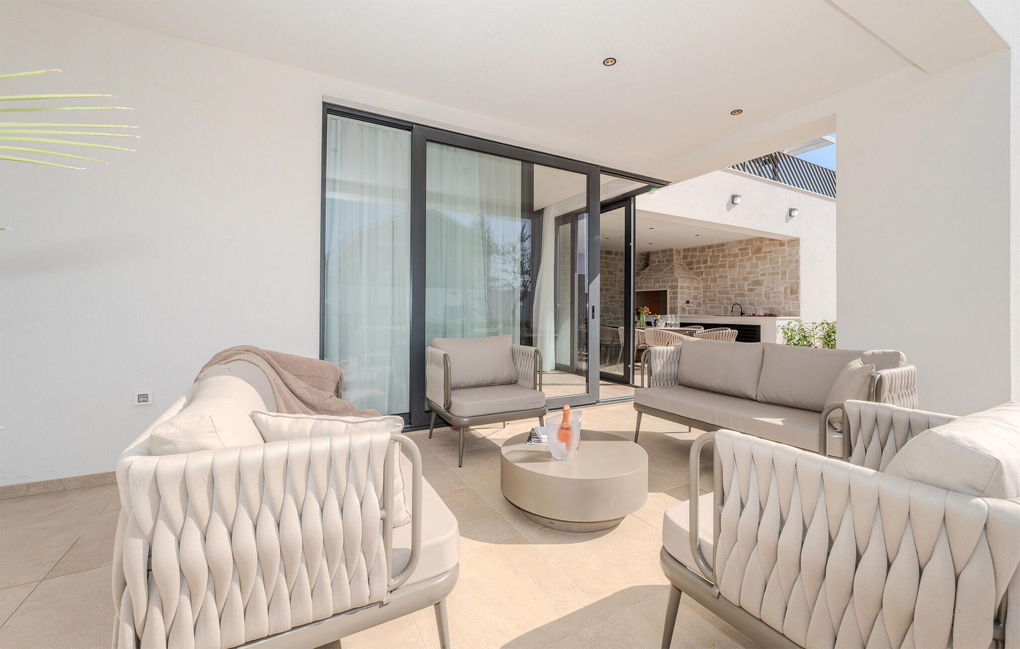 A modern outdoor living area with a glass door leading to a kitchen, comfortable seating, and a stone wall.