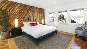 A bedroom with a wooden accent wall, a large bed, a nightstand, a lamp, a potted plant, and a view of the outside through windows. - apt w Balcony, Center of town, Game Room, city view, So Fi stadium, USC, Wi-Fi (Los Angeles)
