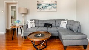 Ein modernes Wohnzimmer mit einem grauen Ecksofa, einem runden Couchtisch aus Holz und einem Gemälde von Wellen an der Wand.