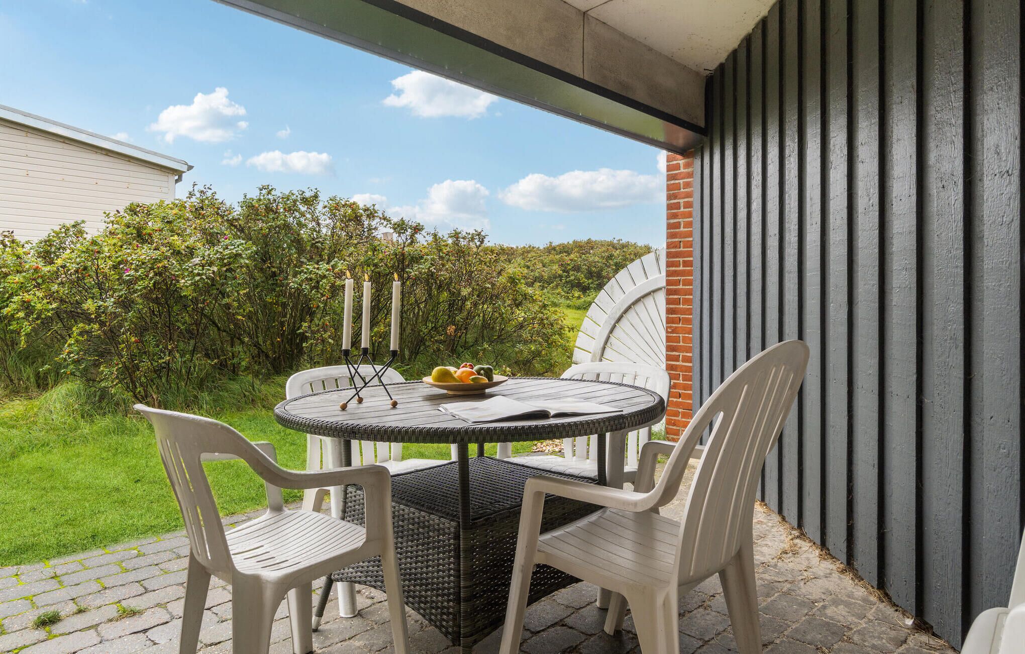Ein runder Tisch mit vier Stühlen, einer Obstschale und drei brennenden Kerzen auf einem Balkon mit Blick auf einen Garten und eine Backsteinmauer.