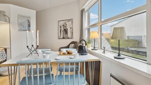 Een eethoek met een tafel gedekt voor twee personen, een waterkoker en uitzicht op een huis door grote ramen.