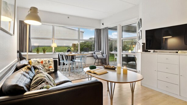 Een moderne woonkamer met een bank, een eettafel, stoelen, een televisie en grote ramen met uitzicht op het omliggende landschap.