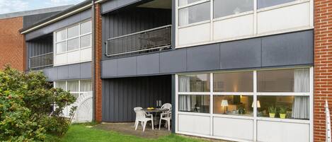 Un immeuble résidentiel de deux étages doté d’un balcon, de grandes fenêtres et d’un petit espace extérieur aménagé pour s’asseoir.