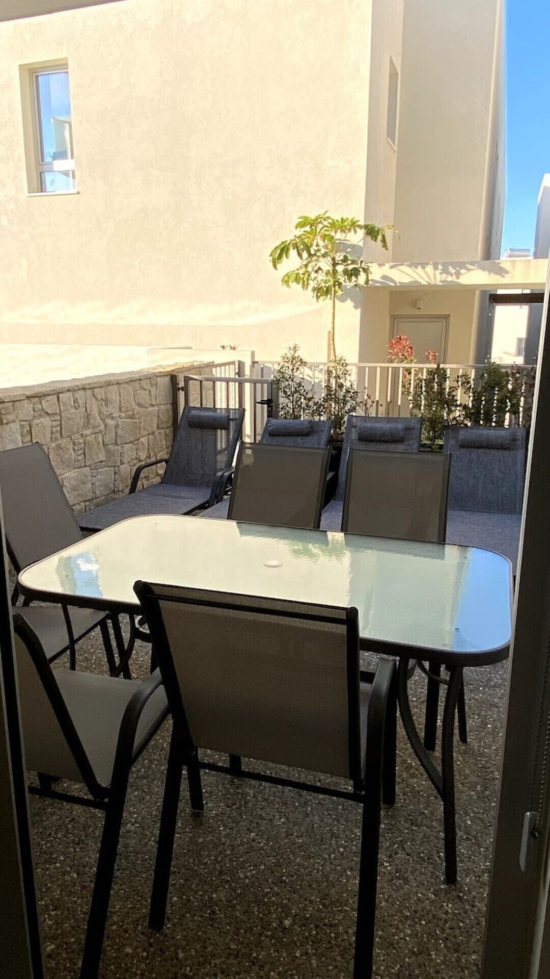 Un patio al aire libre con una mesa de vidrio y sillas negras, rodeado por un muro de piedra y un árbol pequeño.