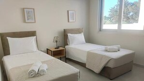 A hotel room with two single beds, a nightstand with a lamp, and a window with a view of trees. - Anchora___ (Paralimni)