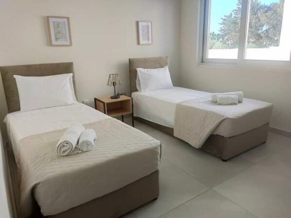 A hotel room with two single beds, a nightstand with a lamp, and a window with a view of trees. - Anchora___ (Paralimni)