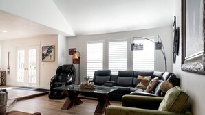 Uma sala de estar com um sofá de couro preto, uma poltrona verde e uma mesa de centro de vidro. Há também uma cadeira de massagem preta, um abajur e um quadro na parede.