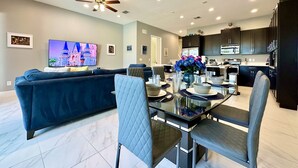 A modern living room with a dark blue sofa, a glass dining table with chairs, a ceiling fan, recessed lighting, and a kitchen area with dark cabinets and appliances. - NEW! 2 Pools + Gym, Kids Water Playground, Near Disney {Chic Retreat} (Davenport)