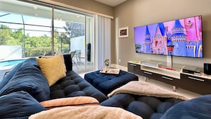 A modern living room with a large flat-screen TV, a sectional sofa, and a dining area with a glass wall overlooking a pool. - NEW! 2 Pools + Gym, Kids Water Playground, Near Disney {Chic Retreat} (Davenport)