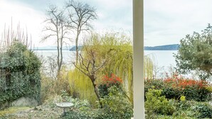 Ein Garten mit verschiedenen Pflanzen und einem Vogelbad, mit Blick auf ein Gewässer und entfernte Berge.