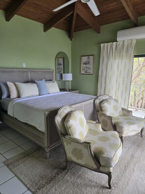 A bedroom with a large bed, two chairs, a lamp, and a window with curtains. - Spectacular View of St Thomas Harbor from both floors & Pool at Bettye’s Villa.  (St Thomas)