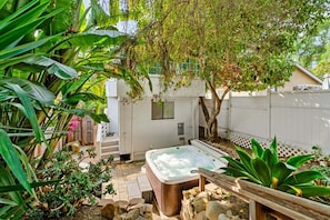 A hot tub in a garden with a wooden deck, surrounded by tropical plants and a white fence.