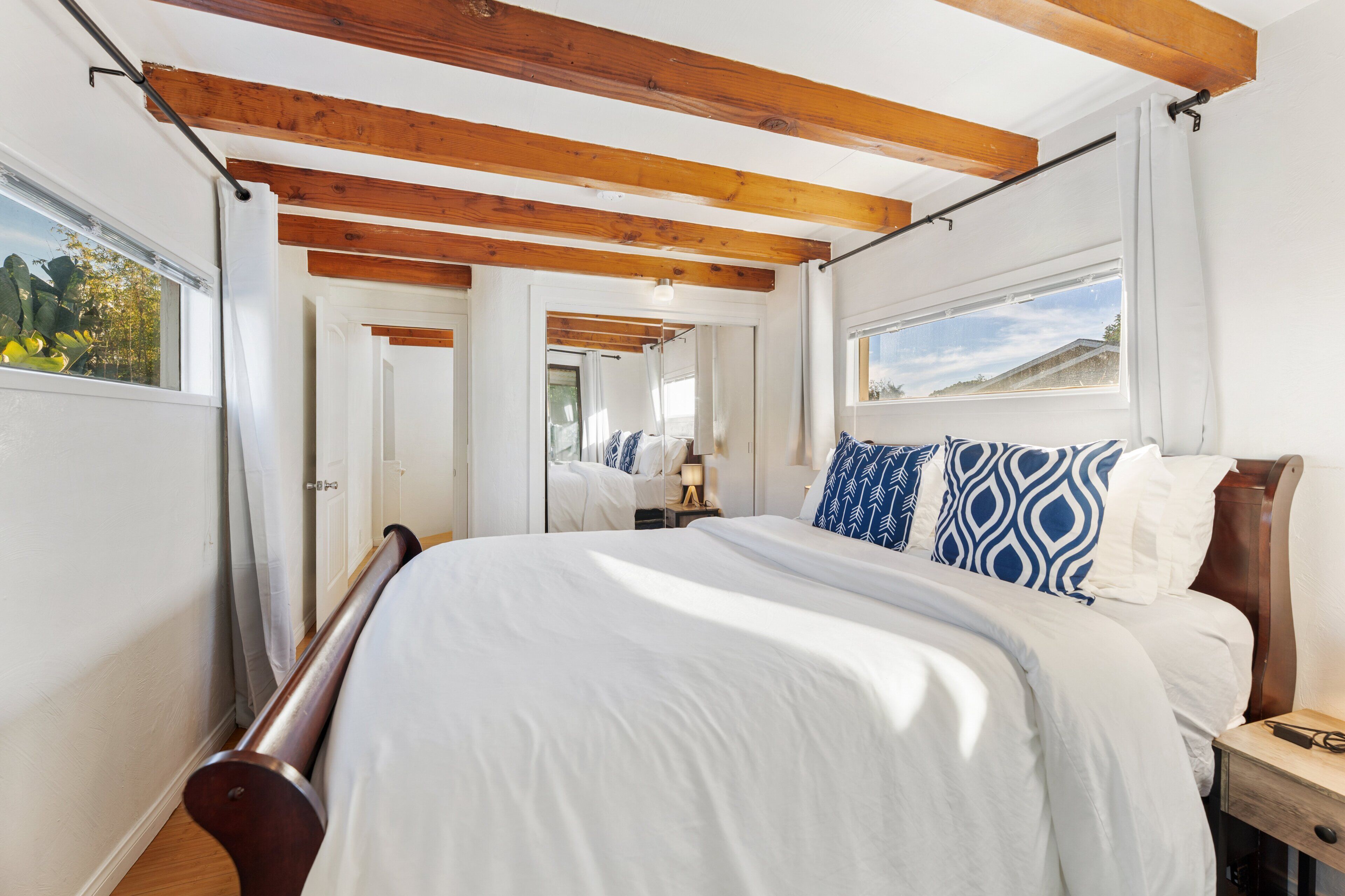 Un dormitorio con techo de madera, cama con ropa de cama blanca y azul, mesita de noche y ventana con vistas.
