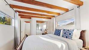 A bedroom with a wooden ceiling, a bed with white and blue bedding, a nightstand, and a window with a view. - 5BR Beach House | Hot Tub, Game Room + Studio (Summerland)