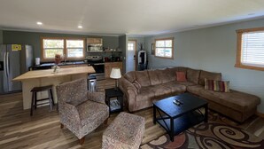 Ein geräumiger Wohnbereich mit einer großen Couch, einem Couchtisch und gemusterten Sesseln. Im Hintergrund ist eine Küche mit Holzschränken und einem Kühlschrank zu sehen.