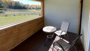 Ein Zimmer mit Holzboden, einem runden Tisch mit zwei Stühlen, Blick auf ein grasbewachsenes Feld und Bäume durch ein großes Fenster und einer Wellblechdecke.