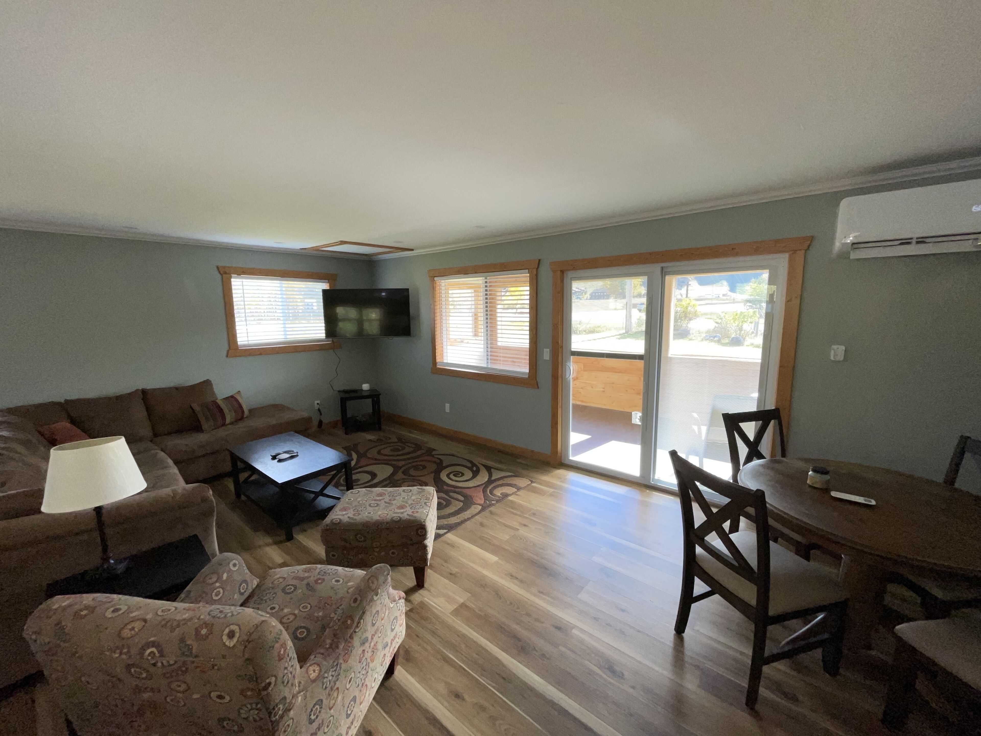 A living room with a sofa, patterned armchairs, a coffee table, and a dining table with chairs. There is a television mounted on the wall and a sliding glass door leading to an outdoor area.