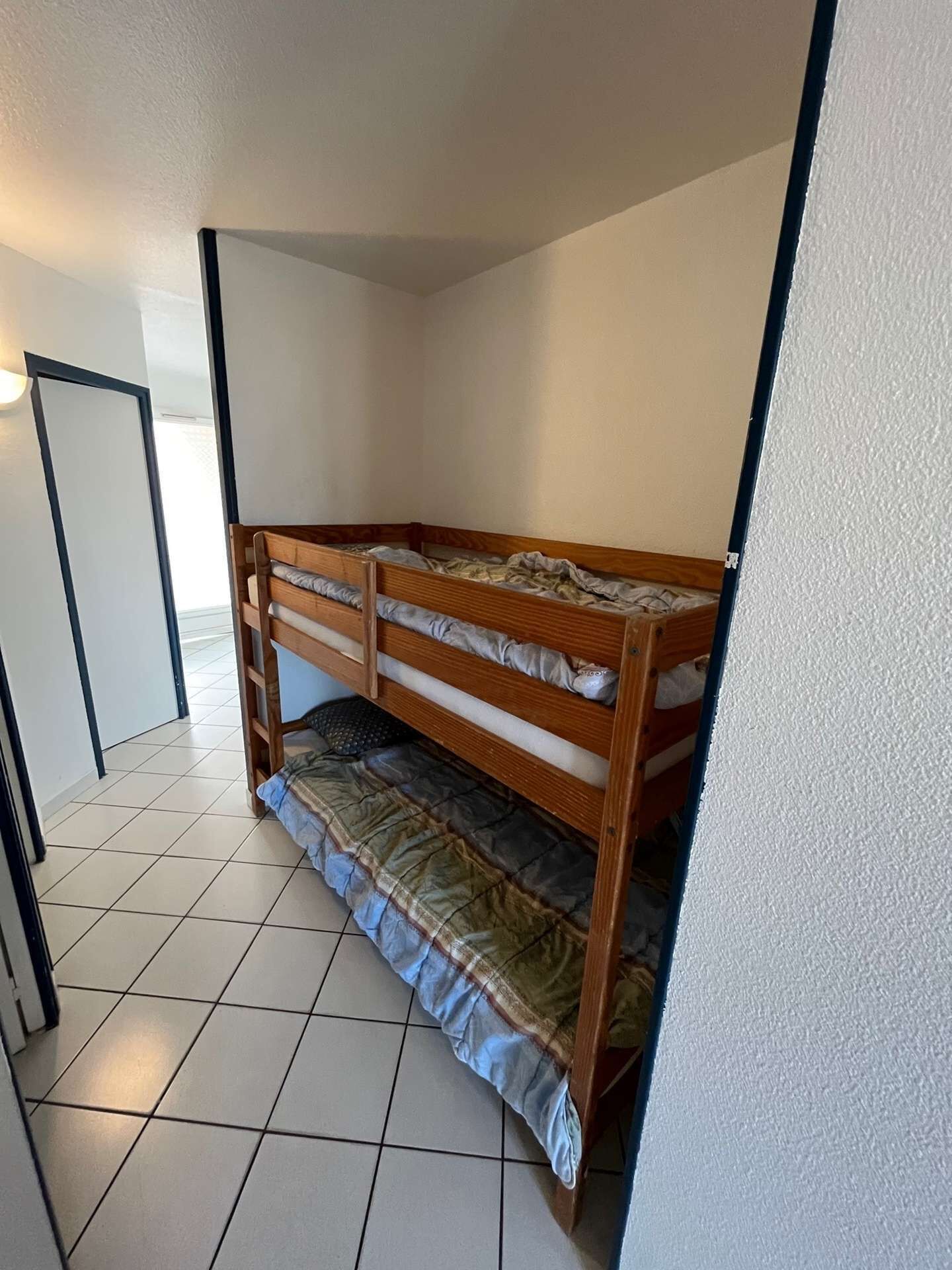 Une petite pièce carrelée, avec un lit superposé, une porte et un luminaire fixé au mur.