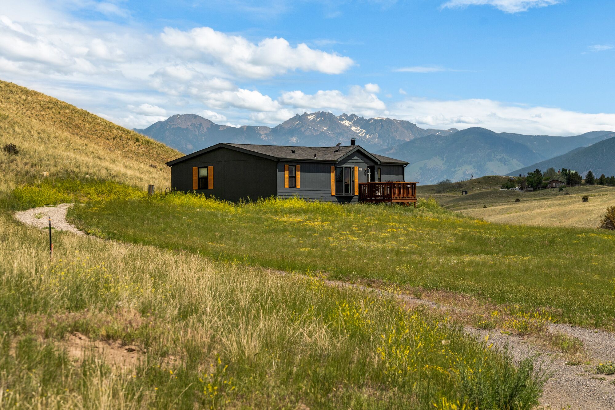 Une maison moderne, de couleur noire, dotée d’une terrasse, entourée d’un champ verdoyant et avec des montagnes en arrière-plan.