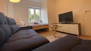 A modern living room with a sofa, a TV on a stand, a small table, and a window with a view of greenery. - Alter Ziegelhof Vechta (Vechta)