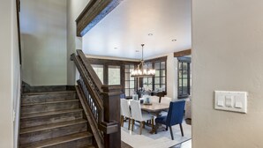 Una escalera conduce a un comedor con candelabro, suelo de madera y ventanas amplias.