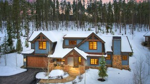 Una cabaña de troncos de dos pisos con cimientos de piedra y techo cubierto de nieve, rodeada por un bosque.